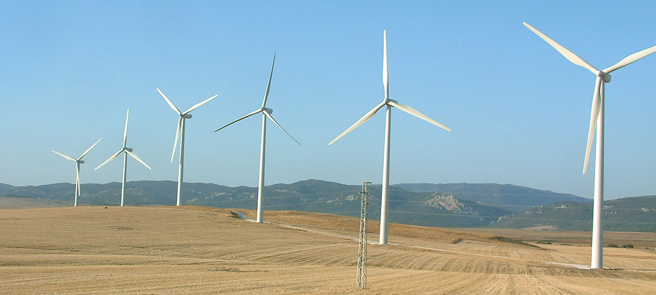 Une rangée de grandes éoliennes blanches se dresse sur un paysage aride sous un ciel bleu clair, chacune d'elles étant soigneusement placée après une évaluation des incidences. Elles sont alignées dans une formation légèrement incurvée, avec des collines ondulantes et des montagnes lointaines ornant l'arrière-plan.