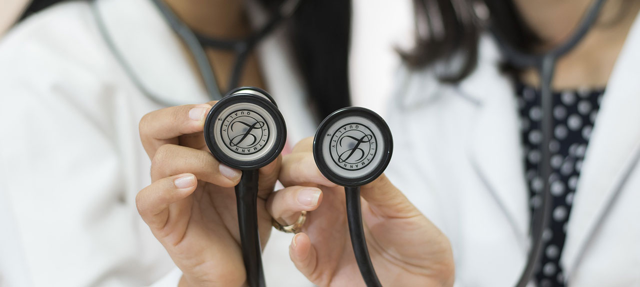 Deux professionnels de la santé en blouse blanche tiennent des stéthoscopes, la cloche tournée vers la caméra. Leurs visages sont invisibles, ce qui met en valeur les stéthoscopes. Cette scène pourrait facilement faire partie d'une publicité pour des services de santé, illustrant l'expertise et la confiance dans le toucher de chaque médecin.