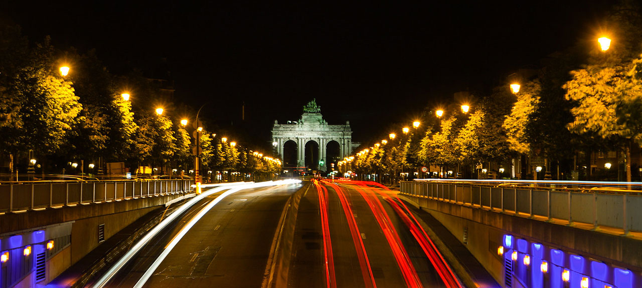 Une scène nocturne d'un monument bien éclairé avec des arbres verts luxuriants et un grand espace à proximité. Des lampadaires lumineux bordent la route, où une longue exposition crée des traînées de lumière blanche et rouge provenant des véhicules qui passent, ajoutant une énergie dynamique au cadre tranquille.