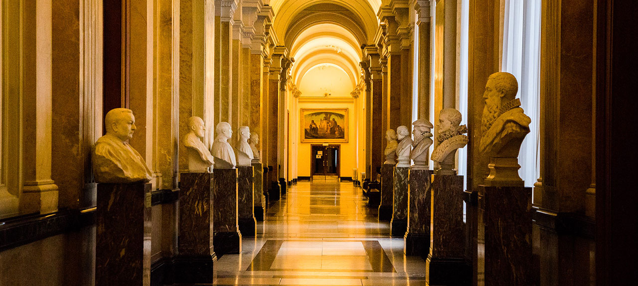 Un grand couloir avec une rangée de bustes en marbre de chaque côté, posés sur de hauts piédestaux. Le couloir présente de hauts plafonds voûtés, des colonnes ornées, des sols polis et des peintures encadrées, créant une impression d'élégance historique.