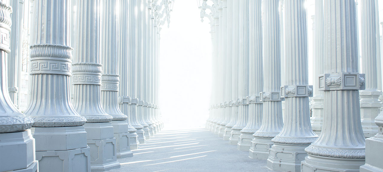 Une série de grands lampadaires blancs décoratifs créent des rangées parallèles menant à une lumière vive à leur extrémité. Les colonnes ont des motifs ornementaux et la scène dégage une ambiance sereine et symétrique.