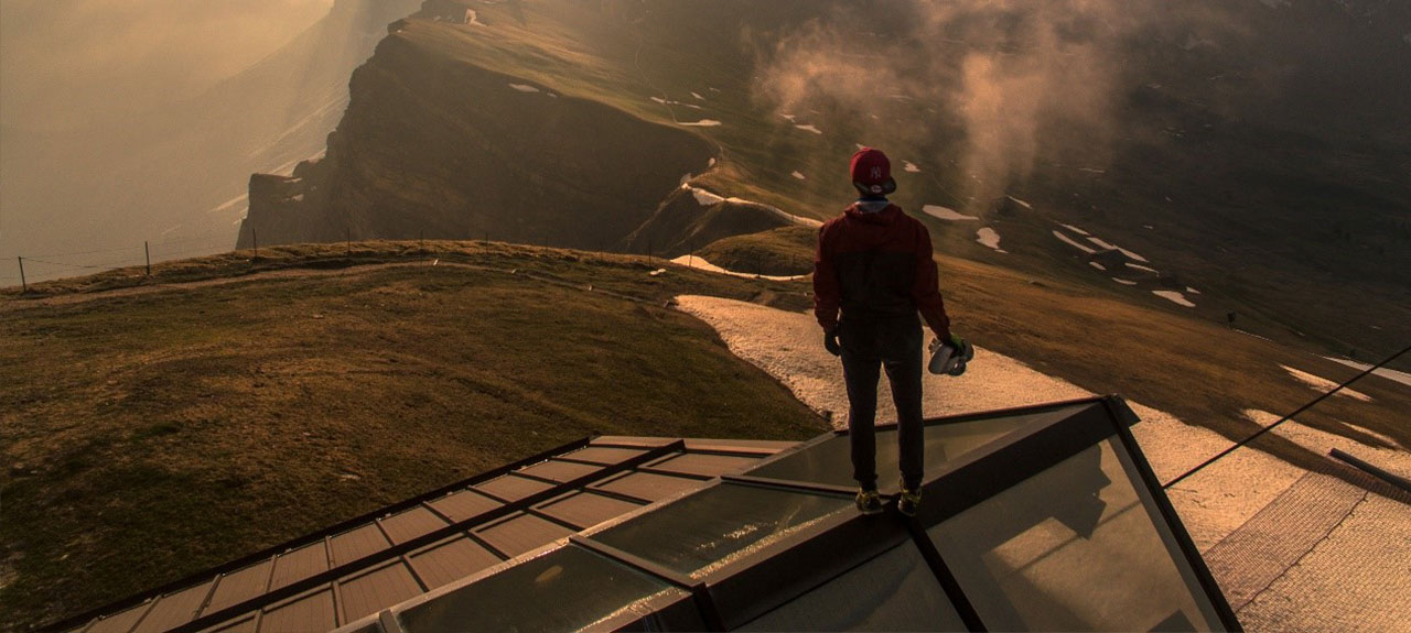 Une personne se tient debout sur un toit en verre, surplombant un vaste paysage de montagnes. Vêtue d'une veste et d'un chapeau rouges, elle tient un casque. Le ciel est partiellement recouvert de nuages et des plaques de neige parsèment le sol. La scène invite à une évaluation sereine des incidents, profondément contemplative dans sa beauté.