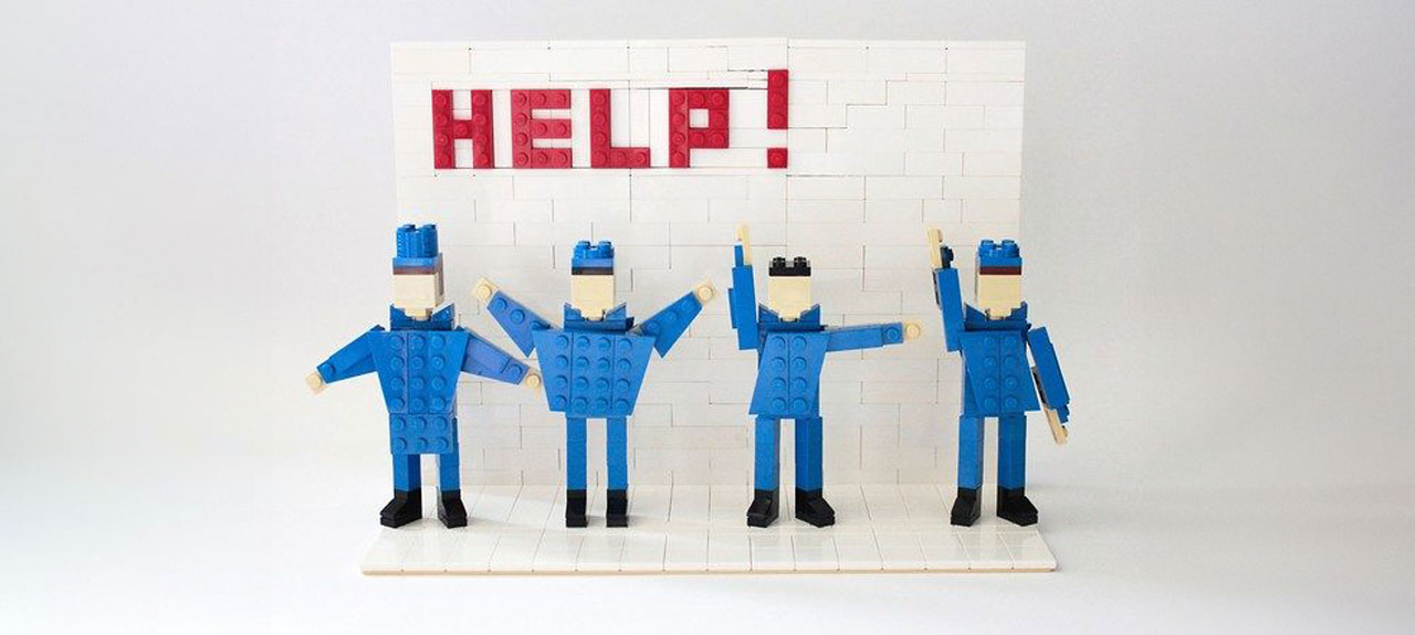 Quatre personnages LEGO en tenue bleue posent devant un mur LEGO blanc avec le mot HELP! écrit en briques rouges. Rappelant la navigation sur les marchés publics, les personnages se tiennent debout sur un socle blanc, chacun adoptant une position de bras unique.