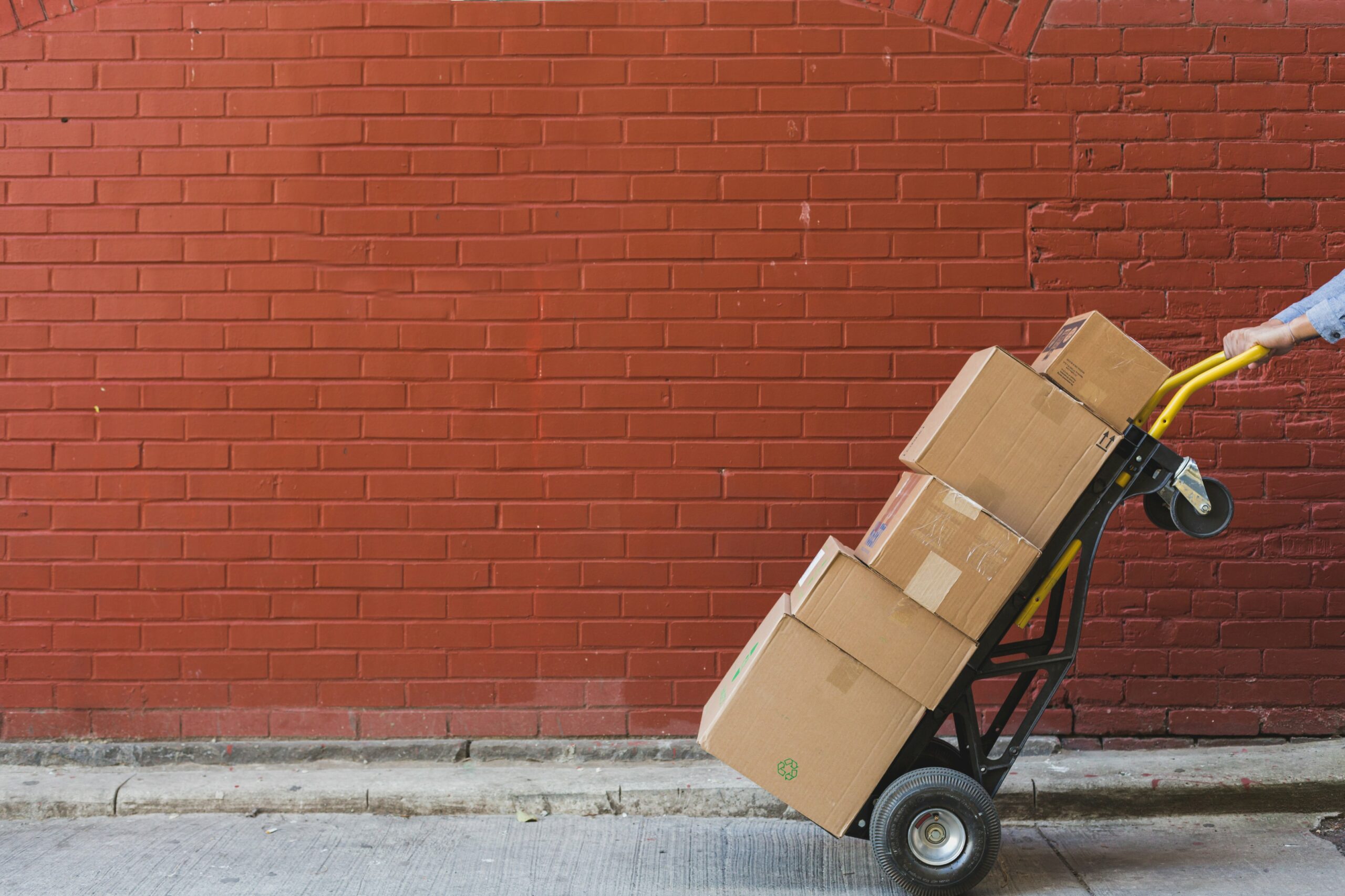 Une personne pousse avec précaution un diable chargé de cartons le long d'un trottoir, démontrant ainsi un contrôle minutieux de la faisabilité des délais. La scène se déroule sur fond d'un mur de briques rouges éclatantes.