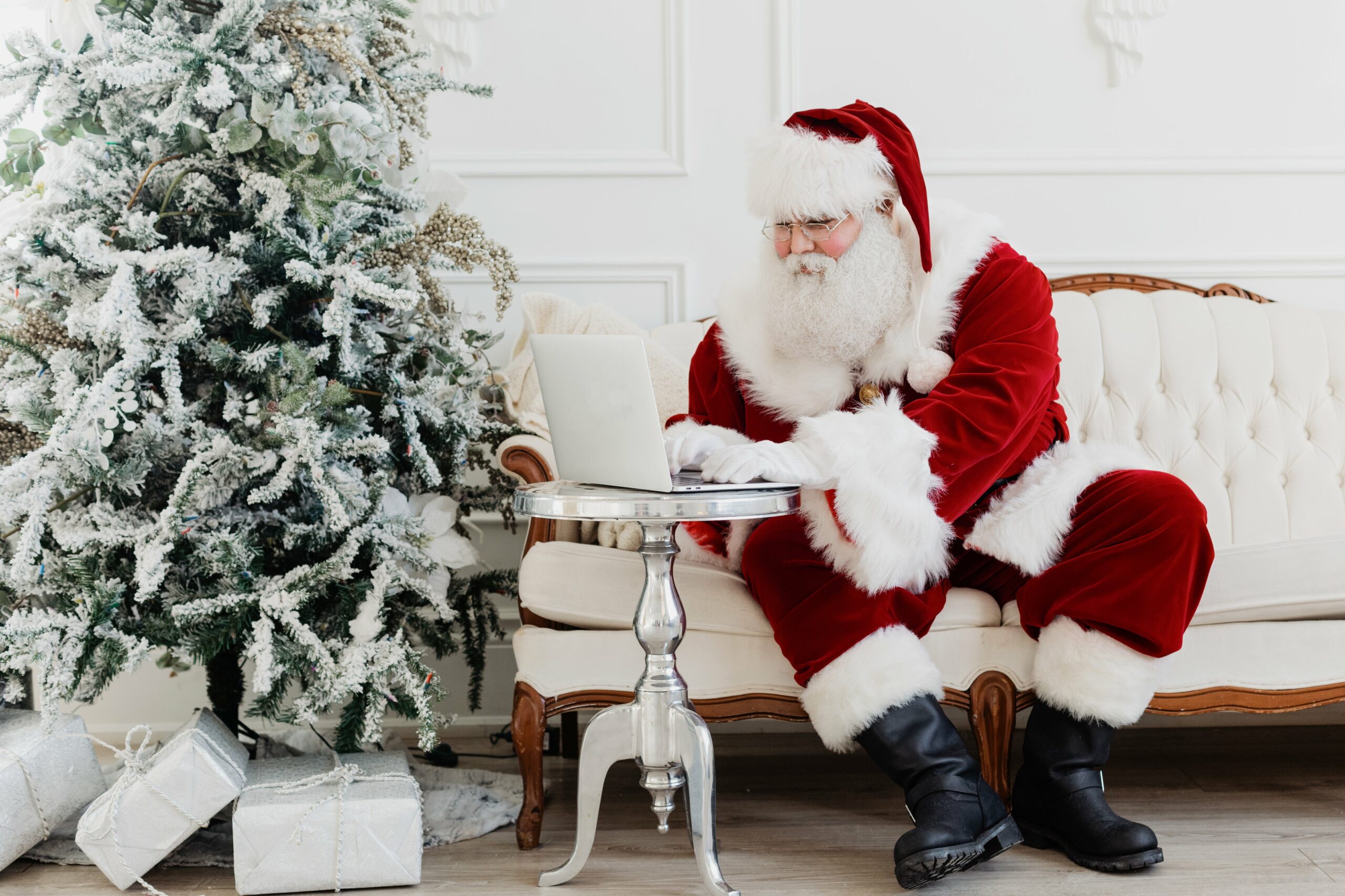 Le Père Noël, vêtu de son costume et de son chapeau rouges emblématiques, est assis sur un canapé blanc et tape sur un ordinateur portable. À proximité, un sapin de Noël givré orné de décorations et de boîtes-cadeaux argentées ajoute de la gaieté à cette élégante pièce. C'est comme s'il veillait à ce que le souhait de chaque enfant soit exaucé en cette période de fêtes.