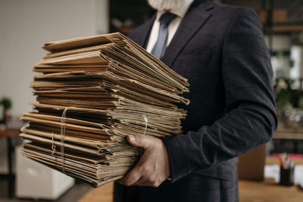 Une personne en costume tient une grande pile de dossiers ou de documents marron attachés dans un bureau, avec des bureaux et des fournitures de bureau visibles en arrière-plan.